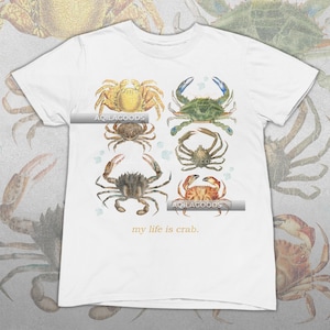 May include: White t-shirt featuring a watercolor design of six different crabs in shades of yellow, green, brown, and orange. The text "my life is crab" is printed in gold at the bottom of the shirt.
