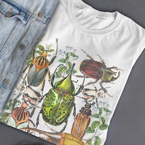 May include: White t-shirt featuring a detailed illustration of various beetles in shades of green, brown, and orange, with sprigs of greenery. The shirt is paired with a light blue denim jacket.