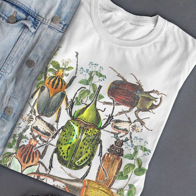 May include: White t-shirt featuring a detailed illustration of various beetles in shades of green, brown, and orange, with sprigs of greenery. The shirt is paired with a light blue denim jacket.
