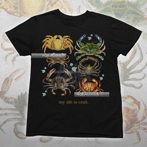 May include: Black t-shirt featuring a watercolor illustration of six different crabs in shades of gold, green, brown, and red. The text "my life is crab" is printed at the bottom. The brand name "AQILAGOODS" is printed on the shirt.