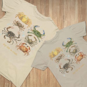 May include: Two beige t-shirts with a watercolor crab design. The crabs are in shades of yellow, green, brown, and orange. The text "my life is crab" is printed on the lower left side of each shirt.