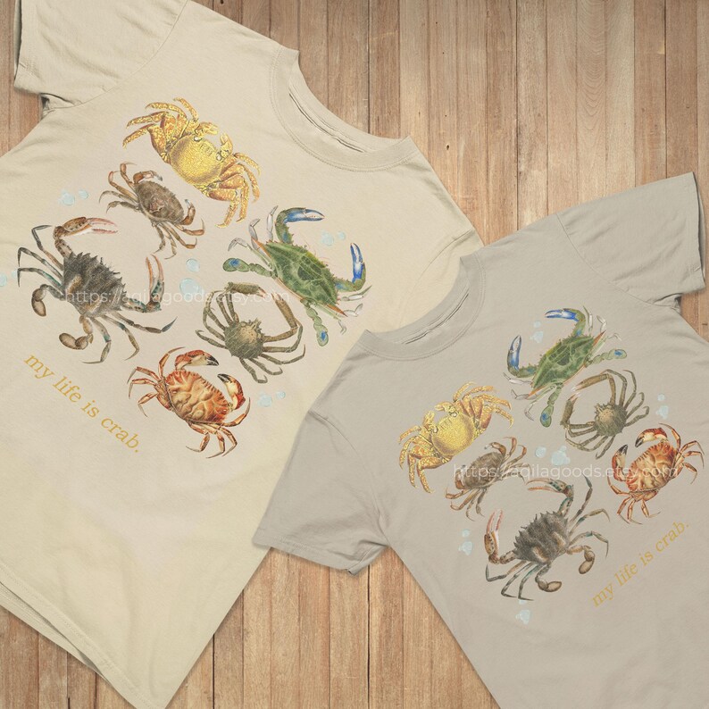 May include: Two beige t-shirts with a watercolor crab design. The crabs are in shades of yellow, green, brown, and orange. The text "my life is crab" is printed on the lower left side of each shirt.