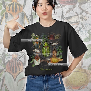 May include: Black t-shirt featuring a detailed illustration of various beetles in shades of green, brown, and white, with botanical elements. The text "The Beetles" is printed below the graphic. The Etsy shop URL is also visible.