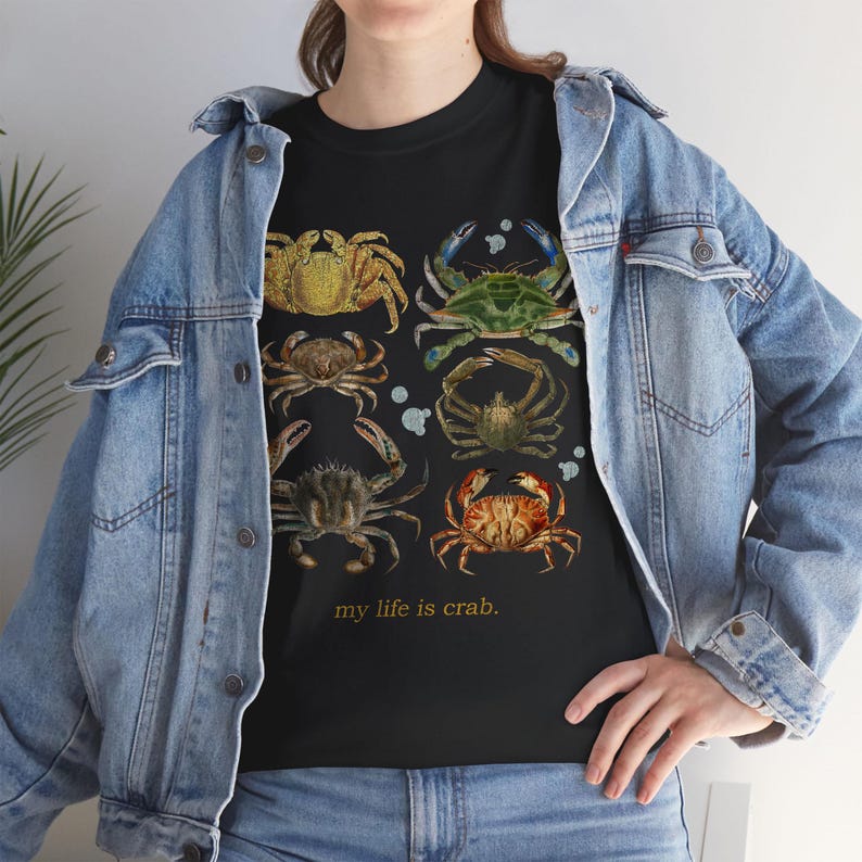 May include: Black t-shirt featuring a collection of illustrated crabs in various colors, including yellow, green, and brown. The phrase "my life is crab" is printed below the crab illustrations. The shirt is worn with a denim jacket.