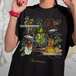 May include: Black t-shirt featuring a detailed illustration of various beetles in shades of green, brown, and yellow, surrounded by botanical elements. The text "The Beetles" is printed at the bottom. The brand name "AQILAGOODS" is also visible.