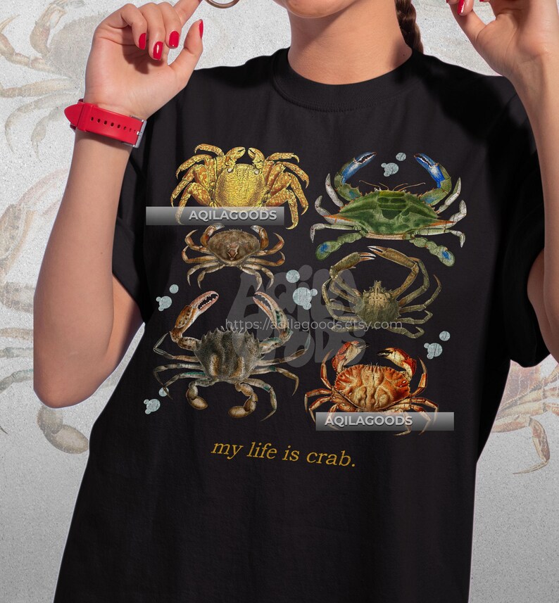 May include: Black t-shirt featuring a collage of six crab illustrations in various colors, including gold, green, and brown. The text "my life is crab" is printed below the images. The shop name "AQILAGOODS" is also visible.
