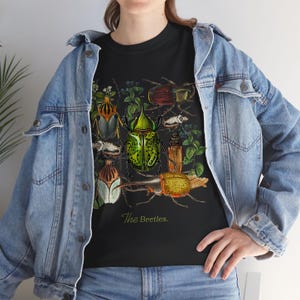 May include: Black t-shirt featuring a detailed illustration of various beetles in vibrant colors. The design includes green, yellow, brown, and red beetles with botanical elements. The text "The Beetles" is printed in a cursive font.
