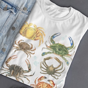 May include: White t-shirt featuring a watercolor illustration of various crabs in shades of yellow, brown, green, and red. The crabs are arranged on the front of the shirt, with a denim jacket partially visible in the background.