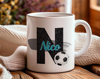Könnte beinhalten: Weiße Keramiktasse mit einem großen, strukturierten schwarzen Buchstaben "N" und dem Namen "Nico" in türkisfarbener Schrift. Eine Fußballgrafik ist ebenfalls vorhanden. Die Tasse steht auf einer Holzoberfläche.