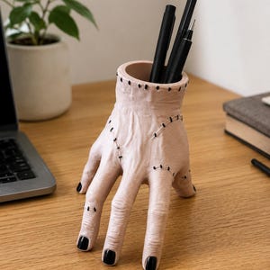 Peut inclure: Un organisateur de bureau en forme de main couleur chair, orné de points de suture noirs sur les doigts et le poignet, avec des ongles peints en noir. Le haut du poignet est ouvert, servant de support à trois stylos noirs verticaux.