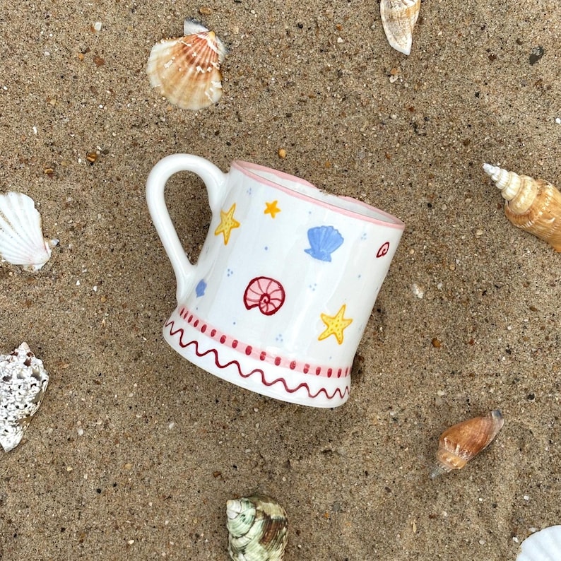 Puede incluir: Taza de cer&aacute;mica blanca con asa, decorada con estrellas amarillas, conchas azules y un dise&ntilde;o en espiral rojo. El borde es rosa y la base tiene un patr&oacute;n rojo ondulado. La taza est&aacute; rodeada de conchas marinas sobre una superficie arenosa.