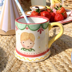 May include: A hand-painted ceramic mug with a yellow handle and a whimsical illustration of a young person. The mug has a green and red border, a wavy yellow base, and a blue and white striped straw. Strawberries and pastries are in the background.