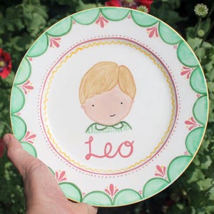 May include: A hand-painted ceramic plate with a cartoon boy illustration. The plate has a white base, a green and pink scalloped border, and the name "Leo" in pink script. The plate is suitable for serving food.
