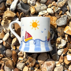 May include: White ceramic mug with a nautical design. The mug features a yellow sun, red and yellow sailboats, blue waves, and blue and white striped accents. The mug has a curved handle.