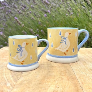 May include: Two ceramic mugs with a yellow background, blue accents, and a white duck illustration. The mugs have blue handles and a blue band at the base. The duck has a blue bow. The background has blue raindrop details.