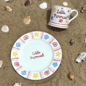 Puede incluir: Un juego de plato y taza de cer&aacute;mica blanca con el tema "La Sirenita". El plato y la taza est&aacute;n decorados con conchas marinas, estrellas de mar y l&iacute;neas onduladas en rosa, azul y amarillo. El juego est&aacute; sobre una superficie arenosa con conchas.