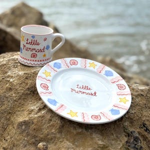 Puede incluir: Un juego de taza y plato de cer&aacute;mica blanca con el tema "Little Mermaid". La taza y el plato est&aacute;n decorados con conchas marinas, estrellas de mar y el texto "Little Mermaid" en rojo. El plato es ovalado.