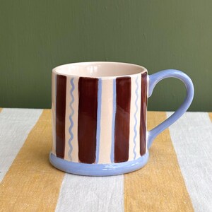 May include: A ceramic mug with a light blue handle and base. The mug features vertical brown and cream stripes, with light blue wavy lines between the stripes. The interior is a light pink color. The mug is on a yellow and white striped surface.