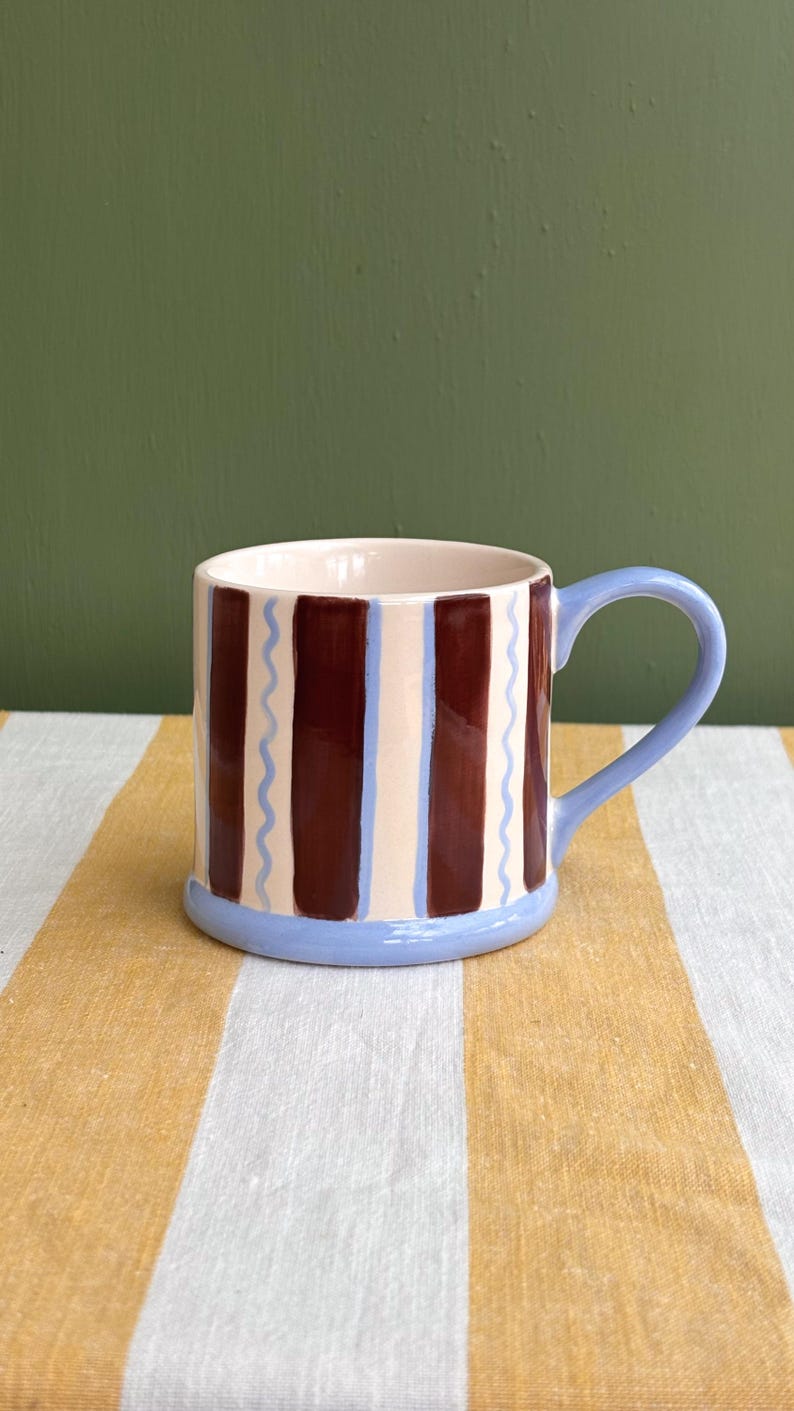 May include: A ceramic mug with a light blue handle and base. The mug features vertical brown and cream stripes, with light blue wavy lines between the stripes. The interior is a light pink color. The mug is on a yellow and white striped surface.