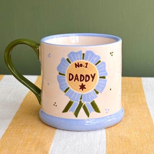 May include: A ceramic mug with a light peach base, a blue rim, and a green handle. The mug features a blue and yellow ribbon design with the text "No.1 DADDY" and a star.