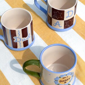 May include: Three ceramic mugs with blue, green, and brown accents. One mug has the word "Dad" in blue script, another has a "No. 1 DADDY" badge, and the third has a checkered pattern with the letters "D", "A", and a star. The mugs have a cream-colored interior.