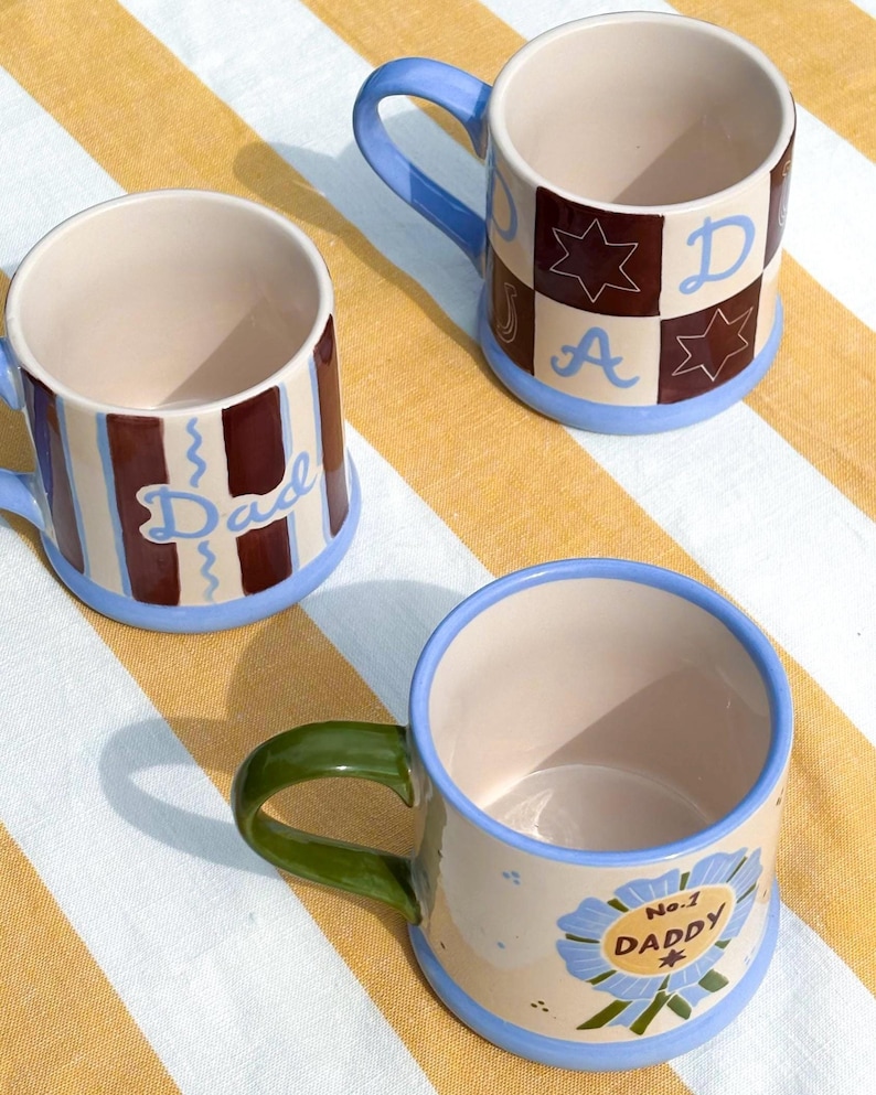 May include: Three ceramic mugs with blue, green, and brown accents. One mug has the word "Dad" in blue script, another has a "No. 1 DADDY" badge, and the third has a checkered pattern with the letters "D", "A", and a star. The mugs have a cream-colored interior.