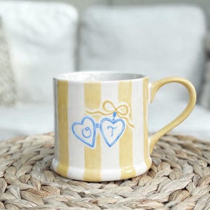 May include: A striped ceramic mug with yellow and white vertical stripes. The mug has two blue heart-shaped designs with the letters "C" and "T". The handle and stripes are yellow. The mug is on a woven mat.