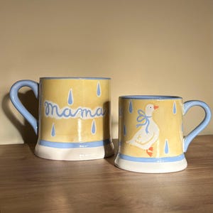 May include: Two ceramic mugs with a yellow background and blue accents. One mug has the word "mama" in cursive, the other features a goose with a blue bow. Both mugs have a raindrop design.
