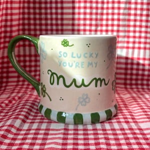 May include: A ceramic mug with a green handle and base stripes. The mug has the text "So lucky you're my mum" in green and light blue. The mug is decorated with shamrocks and small dots.