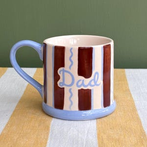 May include: A ceramic mug with a light blue handle and base. The mug features vertical brown and cream stripes, with the word "Dad" in light blue script. Wavy blue lines accent the stripes.