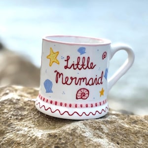 Puede incluir: Taza de cer&aacute;mica blanca con borde y asa rosas, decorada con un dise&ntilde;o ondulado rojo y rosa en la base. La taza presenta las palabras "Little Mermaid" en escritura roja, junto con estrellas de mar amarillas e ilustraciones de conchas azules.