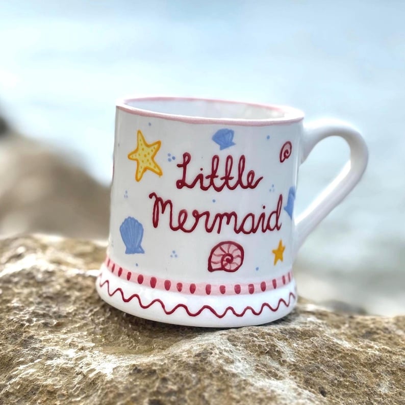 Puede incluir: Taza de cer&aacute;mica blanca con borde y asa rosas, decorada con un dise&ntilde;o ondulado rojo y rosa en la base. La taza presenta las palabras "Little Mermaid" en escritura roja, junto con estrellas de mar amarillas e ilustraciones de conchas azules.