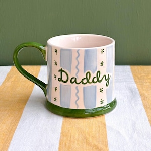 May include: A ceramic mug with a green handle and base, featuring a light blue and white vertical striped pattern. The word Daddy is written in green script on the front, surrounded by small green clover-like accents and a wavy vertical line.