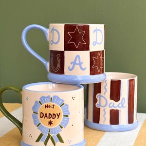 May include: Three ceramic mugs with colorful designs. One mug has a blue handle and a checkered pattern with the word "DAD" and star and horseshoe motifs. Another mug says "No.1 DADDY" on a blue ribbon. The third mug has vertical stripes and the word "Dad".