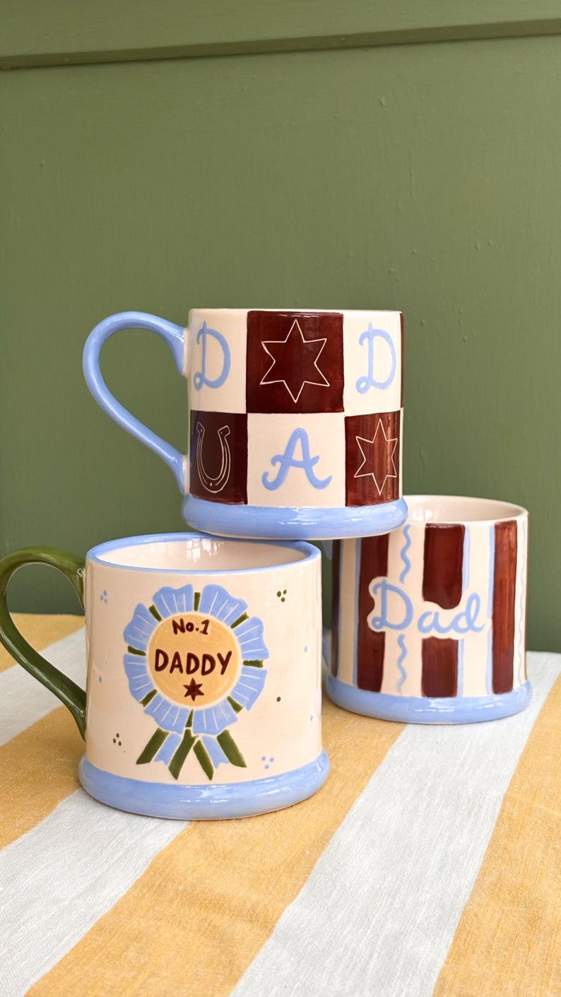 May include: Three ceramic mugs with colorful designs. One mug has a blue handle and a checkered pattern with the word "DAD" and star and horseshoe motifs. Another mug says "No.1 DADDY" on a blue ribbon. The third mug has vertical stripes and the word "Dad".