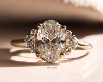 Op de afbeelding: Een gouden ring met een grote, ovaal geslepen diamant als middelpunt. De diamant wordt aan weerszijden geflankeerd door kleinere, ronde diamanten. De ring heeft een delicate band en staat tegen een zachte, neutrale achtergrond. De tekst "Maavi Jewels" is zichtbaar.