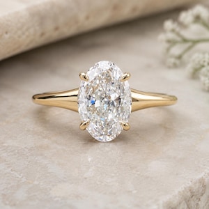 May include: A gold ring featuring a large, oval-cut clear gemstone held in place by four prongs. The band is smooth and tapers towards the centre setting, showcasing a polished yellow gold finish that contrasts with the faceted, brilliant-cut stone.