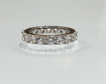 May include: A silver ring with a continuous row of clear, round-cut gemstones. The ring is set with multiple small, sparkling stones, creating a full eternity band design. The ring is displayed on a reflective surface.