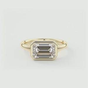 Puede incluir: Un anillo de oro con engaste de bisel que presenta un diamante talla esmeralda transparente.
