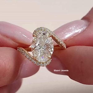 Op de afbeelding: Een gouden verlovingsring met een grote, ovaal geslepen diamant. De ring heeft een uniek ontwerp met een wervelende band bezet met kleinere diamanten. De ring wordt vastgehouden door de vingers van een persoon. De tekst "Maavi Jewels" is zichtbaar.