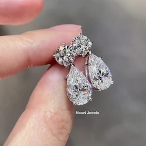 May include: Sparkling diamond earrings with a cluster of round-cut stones and a pear-shaped drop. The earrings are held up by a hand, with the brand name "Maavi Jewels" visible. A luxurious jewellery item.