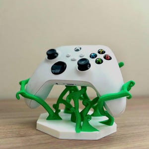 Può includere: Controller Xbox bianco su un supporto personalizzato. Il supporto ha una base esagonale bianca con supporti verdi a forma di vite. Il controller ha levette nere e pulsanti di azione colorati.