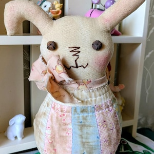 May include: A handmade fabric bunny doll with button eyes and stitched facial features. The bunny wears a pink floral scarf and a patchwork skirt in pastel colors. A small white bunny figurine is visible in the background.