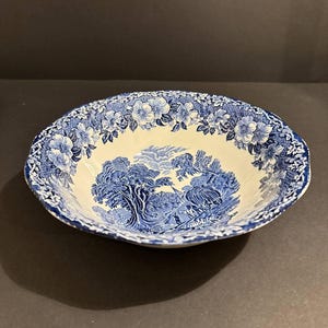 Könnte beinhalten: Eine runde, blau-weiße Keramikschale mit Landschaftsdesign. Die Schale hat ein detailliertes blaues Transfermuster aus Bäumen, Figuren und einem floralen Rand. Die Schale hat einen Durchmesser von etwa 25 cm.