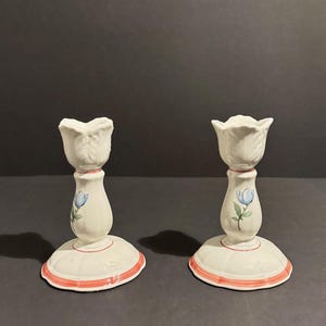 Könnte beinhalten: Zwei weiße Keramik-Kerzenhalter. Jeder hat eine tulpenförmige Spitze, eine bemalte blaue Blume und einen roten Rand. Die Kerzenhalter stehen auf einem dunklen Hintergrund.