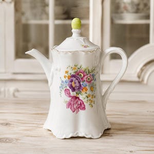 Può includere: Teiera in porcellana bianca con un design floreale con fiori viola, rosa e blu. La teiera ha un manico curvo, un beccuccio e un coperchio con un pomello verde. La teiera è decorata con finiture dorate e poggia su una superficie di legno chiaro.