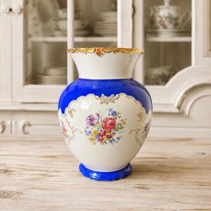 Könnte beinhalten: Eine Porzellanvase mit einem breiten, gewellten Rand und Goldverzierung. Der Körper ist in einen tiefblauen oberen Bereich und einen cremefarbenen unteren Bereich unterteilt, verziert mit einem zentralen Blumenstrauß in Rot, Gelb und Blau.
