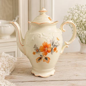 Puede incluir: Tetera de porcelana color crema con tapa, con diseños florales y detalles en naranja. La tetera tiene un pico y un asa curvados, y está sobre una superficie de madera. El patrón floral incluye flores y hojas naranjas y grises.