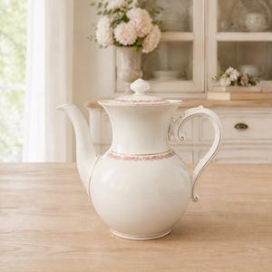 Könnte beinhalten: Eine weiße Porzellan-Kaffeekanne mit gebogenem Ausguss und kunstvollem Griff. Die Kanne hat ein rosa Blumenband und goldfarbene Verzierungen. Der Deckel hat einen kleinen Knauf. Die Kanne steht auf einer hellen Holzoberfläche.