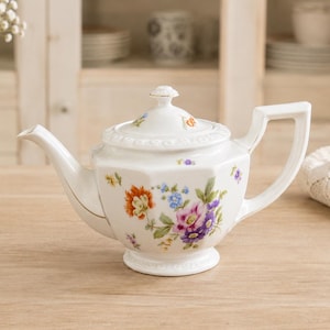 Könnte beinhalten: Weiße Porzellan-Teekanne mit floralem Design mit orangefarbenen, lila und blauen Blumen. Die Teekanne hat einen goldenen Rand, einen Deckel mit kleinem Knauf und einen quadratischen Griff. Sie steht auf einer Holzoberfläche.